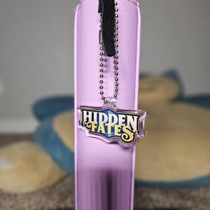 May include: A vertical, light purple card binder with a black leather strap and a silver chain. The binder has a "HIDDEN FATES" logo charm attached. The background is blurred, with a blue and yellow figure visible.