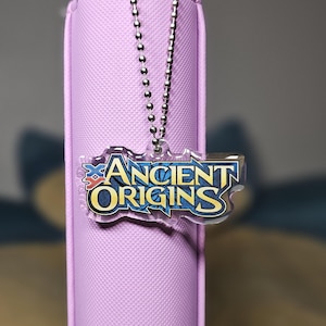 May include: A light purple pencil case with a silver ball chain and a clear acrylic charm. The charm has the words "ANCIENT ORIGINS" in blue and gold lettering.