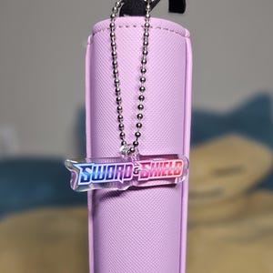 May include: A lavender zippered pouch with a silver chain attached to a clear plastic charm with the words "Sword & Shield" printed in blue and pink.