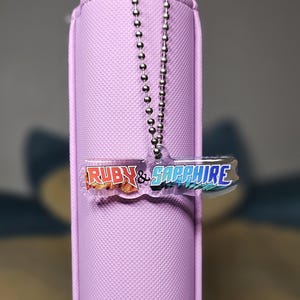 May include: A lavender-colored case with a silver ball chain and a clear acrylic charm. The charm reads "RUBY & SAPPHIRE" in red and blue lettering. The case has a textured surface.