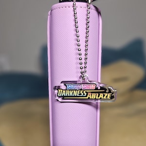 May include: A pink zippered pouch with a silver chain attached to a clear plastic charm with the text "SWORD & SHIELD DARKNESS ABLAZE" in rainbow colors.