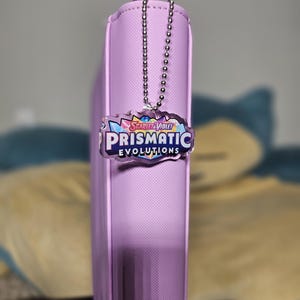 May include: A purple binder with a silver chain attached to a clear plastic charm. The charm has a colorful graphic with the text "Scarlet & Violet Prismatic Evolutions".