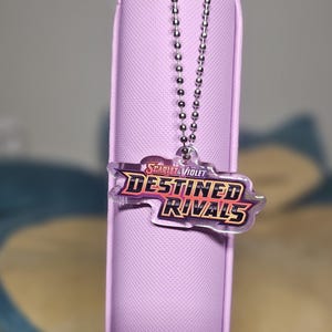 May include: A purple zippered pouch with a silver chain attached to a clear plastic charm with the text "Scarlet & Violet Destined Rivals" in a yellow and orange font.
