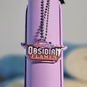 May include: A purple zippered pouch with a silver chain attached to a clear plastic charm with the text "Scarlet & Violet Obsidian Flames" in a colorful, stylized font.
