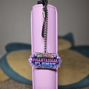 May include: A light purple card binder with a black zipper pull and a silver ball chain. A clear acrylic charm with the text "MEGA EVOLUTION PHANTASMAL FLAMES" hangs from the chain.