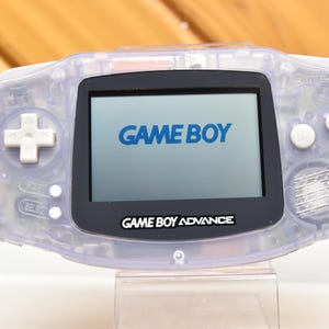 May include: A transparent Game Boy Advance handheld console. The screen displays "GAME BOY" in blue text. The console has a directional pad, buttons, and the text "GAME BOY ADVANCE" below the screen.