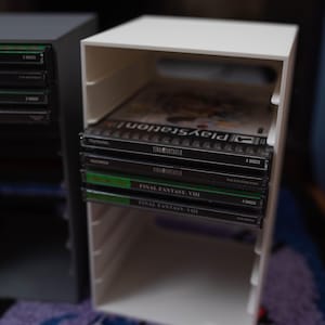 Puede incluir: Unidad de almacenamiento de juegos rectangular blanca que contiene varias carcasas de juegos de PlayStation. Las carcasas son negras con texto verde y blanco, incluyendo "FINAL FANTASY VIII" y "4 DISCS". Otra unidad similar es visible.