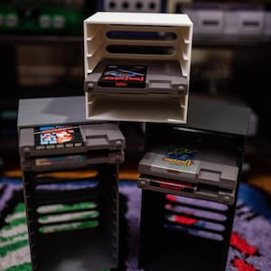 May include: Three retro video game cartridge holders in white, grey, and black, each holding a game cartridge. The cartridges are labelled with game titles. The holders are on a colourful rug.