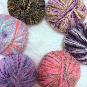 May include: A selection of seven colourful yarn balls, featuring hues of purple, pink, blue, and green. The yarn is arranged in a circle against a white, fluffy backdrop. The yarn has a soft, textured appearance, ideal for knitting or crochet.