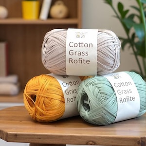 May include: Three skeins of yarn in beige, yellow-orange, and light green are stacked on a wooden table. Each skein has a white label with the text "Cotton Grass Rofite". The yarn is tightly wound and appears to be made of a natural fiber.