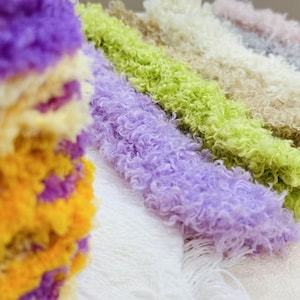 Puede incluir: Primer plano de hilos de lana coloridos, incluyendo morado, verde y beige. Los hilos tienen un aspecto rizado y texturizado, ideal para manualidades. La imagen resalta los colores vibrantes y la textura suave de la lana, adecuada para diversos proyectos.