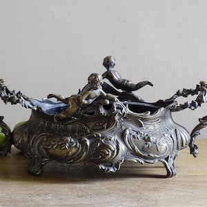 Könnte beinhalten: Antike bronzene Mittelstückschale mit Cherubfiguren. Die Schale hat ein detailliertes Blumen- und Scroll-Design mit Griffen und Füßen. Die Cherubim befinden sich in liegender Pose. Die Schale steht auf einer Holzoberfläche.