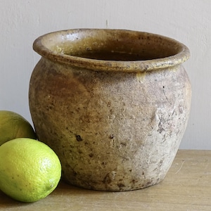 Może przedstawiać: Zniszczony, okrągły, beżowy ceramiczny garnek z szerokim otworem, ustawiony na drewnianej powierzchni. Trzy zielone limonki są ułożone przed garnkiem. Garnek ma teksturowaną powierzchnię i lekko nierówny brzeg.