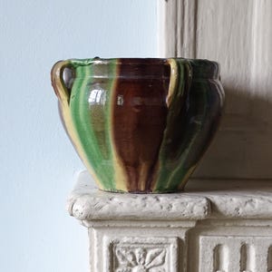 May include: A round ceramic bowl with a glossy finish, featuring vertical stripes of dark brown, forest green, and mustard yellow. The bowl has a wide opening, a tapered base, and two small, curved handles attached to the upper rim on opposite sides.