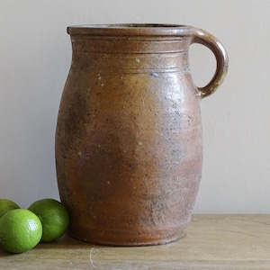 Pode incluir: Uma jarra grande e antiga de grés castanho com uma pega, provavelmente usada para armazenar líquidos. A jarra tem uma base larga e um pescoço mais estreito. Três limas verdes estão em frente à jarra numa superfície de madeira.