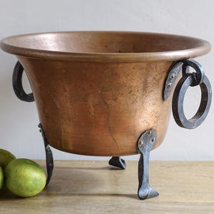 Peut inclure: Un chaudron en cuivre avec un large bord et des poignées et pieds en fer riveté. Le chaudron a une forme arrondie et une couleur brun-rougeâtre chaude. Trois citrons verts sont devant le chaudron.