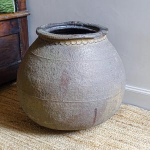 May include: A large, round, antique ceramic pot with a textured, light gray surface. The rim is dark gray with a scalloped edge. The pot has horizontal lines and reddish-brown stains. It sits on a woven rug.