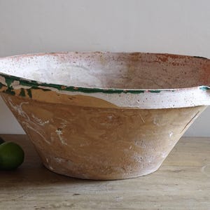 May include: A large, conical earthenware mixing bowl with a wide, flat rim. The exterior is a natural tan color, while the interior features a white glaze with a green painted band along the top edge and a small pouring spout on the side.
