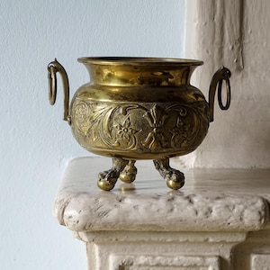Op de afbeelding: Een messing kom met een reliëf bloemmotief, twee zijhandgrepen met hangende ringen en drie pootjes in de vorm van dierenpoten. Het metalen oppervlak heeft een gepolijste goudkleurige afwerking met donkere patina-accenten in de verdiepingen.