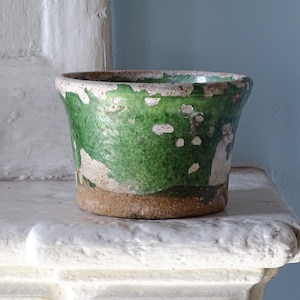 Pode incluir: Um pequeno vaso de cerâmica redondo com um esmalte verde desgastado e manchas brancas visíveis de argila exposta. A base não é esmaltada, exibindo um tom terroso natural. A borda é ligeiramente alargada e a textura é irregular.