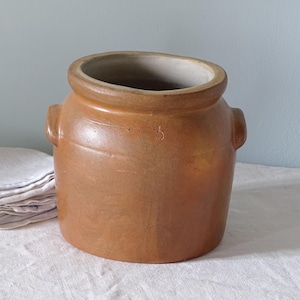 May include: A cylindrical tan stoneware crock featuring a thick rolled rim, two small side handles, and a stamped number 3 on the front. The interior is a light grey color, and the exterior has a smooth, glazed finish with subtle color variations.