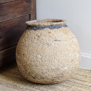 May include: A large, textured, beige decorative vase with a wide, rounded body and a dark rim. The vase sits on a woven jute rug next to a wooden cabinet. The vase has a rustic, aged appearance, suggesting it is an antique.
