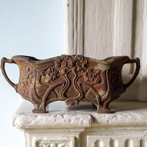 Puede incluir: Una jardinera rectangular de hierro fundido con un acabado marrón desgastado, que presenta motivos florales en relieve y patrones de volutas. El recipiente tiene dos asas laterales, cuatro patas cortas y una superficie texturizada con oxidación.
