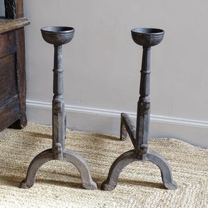 Pode incluir: Dois castiçais antigos de metal cinza escuro. Cada um tem uma tigela redonda no topo, uma coluna decorativa e uma base de três pernas. Os castiçais estão em um tapete tecido.
