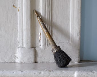 May include: A vintage paint brush featuring a weathered, light brown wooden handle with a small circular hole at the top and a metal ferrule securing a dense cluster of dark, coarse bristles. The tool shows signs of age and past use.