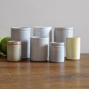 May include: A selection of ceramic jars in different sizes and colours, including grey, tan, and yellow. The jars are cylindrical with lids, and are arranged on a wooden surface. Limes are visible in the background.