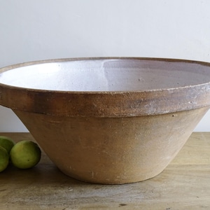 May include: A large, antique brown mixing bowl with a white interior and a pouring spout. The bowl has a wide rim and a conical shape. Three green fruits sit to the left of the bowl on a wooden surface. The bowl is likely made of stoneware.