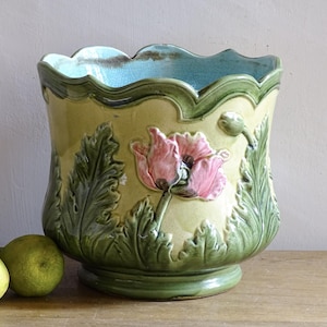 Op de afbeelding: Een decoratieve keramische plantenbak met een geschulpte rand en een voet. De plantenbak heeft een verhoogd bloemmotief met roze klaprozen en groene bladeren tegen een gele achtergrond. De binnenkant is lichtblauw.