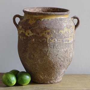 Può includere: Un grande vaso in terracotta usurata con due manici laterali, caratterizzato da una superficie marrone ruvida e testurizzata e da una fascia di smalto giallo attorno al bordo superiore. Tre lime verde brillante sono posizionati davanti alla base.