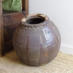Op de afbeelding: Een grote, ronde, antieke bruine keramische pot met een brede opening en een getextureerd oppervlak. De pot heeft een decoratieve geschulpte rand en verticale en horizontale lijnen die een rasterpatroon vormen.