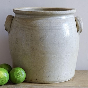 May include: A large, off-white ceramic crock featuring a wide mouth, a thick rim, and two side handles with vertical ribbed detailing. The exterior surface displays a weathered, speckled texture with subtle horizontal incised lines near the top.