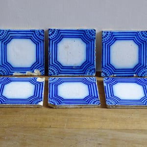 Könnte beinhalten: Sechs antike quadratische Fliesen mit weißer Mitte und blauem geometrischen Rand. Die Fliesen sind auf einer Holzoberfläche angeordnet. Der blaue Rand weist Linien und gepunktete Muster auf. Einige Fliesen weisen Gebrauchsspuren auf.