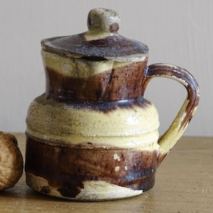 Op de afbeelding: Een keramische kan met deksel, voorzien van een tweekleurig glazuur in donkerbruine en lichtgele tinten. De kan heeft een ronde vorm met horizontale ribbels, een gebogen handvat en een bijpassend deksel met een kleine knop.