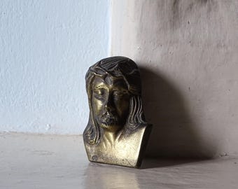 Könnte beinhalten: Eine metallene Wandplakette in Messingoptik mit einem skulpturalen Relief eines Gesichts mit geschlossenen Augen und langem, welligem Haar. Die Figur trägt eine Dornenkrone auf der Stirn, mit detaillierten Gesichtszügen und glatter Oberfläche.