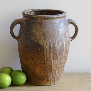 May include: A large, rustic brown ceramic vase with two handles. The vase has a textured surface with dripped glaze effects. Three green limes sit in front of the vase on a wooden surface.