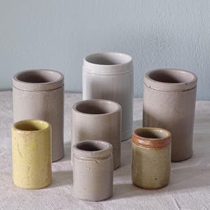 May include: A collection of seven cylindrical ceramic crocks in various sizes and colours, including muted grey, bright yellow, and earthy brown tones. Each vessel features a simple, straight-sided design with a distinct rim and a smooth finish.