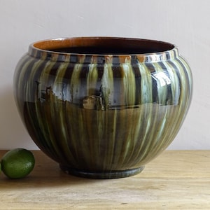May include: A large, round ceramic planter with a glossy finish. The planter features vertical stripes in shades of green, brown, and black. Three green limes sit on a wooden surface in front of the planter. The planter is approximately 30 cm in diameter.