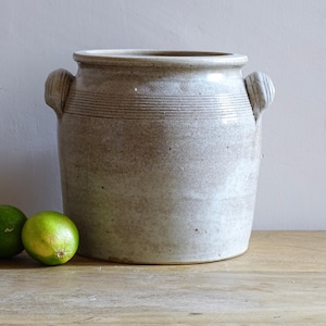 Könnte beinhalten: Ein großer, cremefarbener Steinguttopf mit zwei Griffen und horizontalen, geätzten Linien. Drei grüne Limetten liegen vor dem Topf auf einer Holzoberfläche. Der Topf hat eine rustikale, Vintage-Ästhetik.