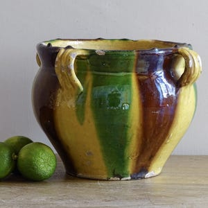 Può includere: Un vaso in ceramica rotondo con due manici laterali, caratterizzato da una smaltatura lucida a strisce verticali gialle, verdi e marrone scuro. Il bordo presenta una forma smerlata con segni di usura. Tre lime verdi sono poste a sinistra.