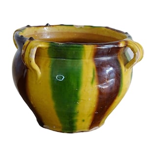 Op de afbeelding: Een gele, groene en bruine gestreepte keramische pot met twee handvatten. De pot heeft een ronde vorm en een licht uitlopende rand.