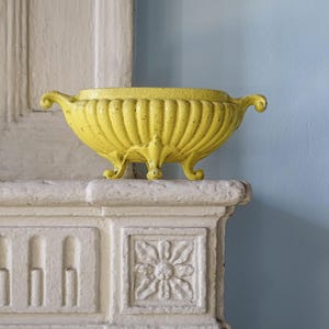 Könnte beinhalten: Eine leuchtend gelbe, ovale Metallschale mit einem gerippten, kannelierten Außendesign. Die Schale steht auf vier kleinen, geschwungenen Füßen und verfügt über zwei kleine Griffe an den gegenüberliegenden Seiten mit einer strukturierten Lackierung.