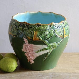 May include: A green ceramic planter with a light blue interior and a scalloped rim. The planter features a raised relief design of leaves and a pink and white flower. Three green limes sit in front of the planter.
