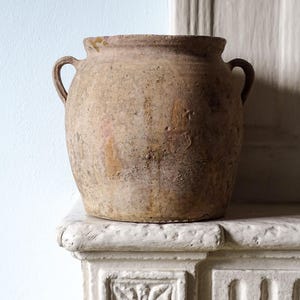 Peut inclure: Un pot en terre cuite rustique et patiné avec une large ouverture circulaire et deux petites anses latérales incurvées. La surface extérieure présente une finition tachetée dans des tons terreux de brun et de beige avec des signes d'usure.