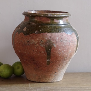 Può includere: Un recipiente rustico in terracotta con un bordo largo e svasato e un corpo bulboso. La parte superiore presenta una finitura smaltata verde scuro gocciolante, mentre la sezione inferiore rimane non smaltata, mostrando una superficie in argilla.