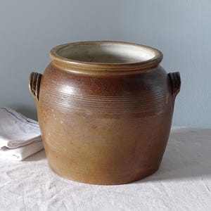 Op de afbeelding: Een bruine stenen pot met een wijde opening, een dikke rand en twee handvatten aan de zijkanten. De buitenkant heeft een kleurverloop van donkerbruin aan de bovenkant naar een lichtere tint bruin aan de basis, met horizontale lijnen.