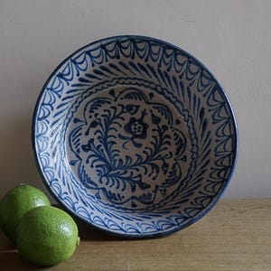 May include: A large, round ceramic bowl with a blue and white floral design. The bowl has a wide rim and is decorated with intricate patterns. Three green limes are arranged in front of the bowl on a wooden surface.